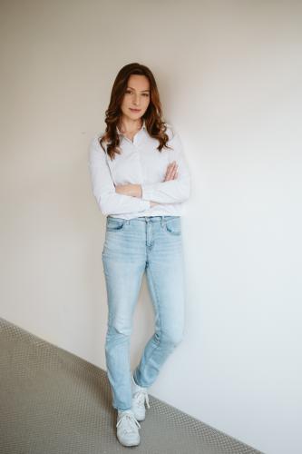 Portrét ženy s dlouhými vlasy v bílé košili a modrých džínách opírá se o stěnu