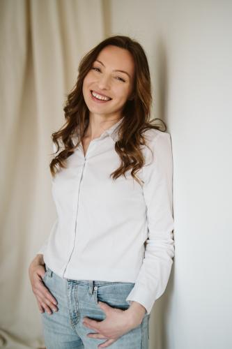Smějící se žena s dlouhými vlasy v bílé košili a modrých džínách opřená o stěnu